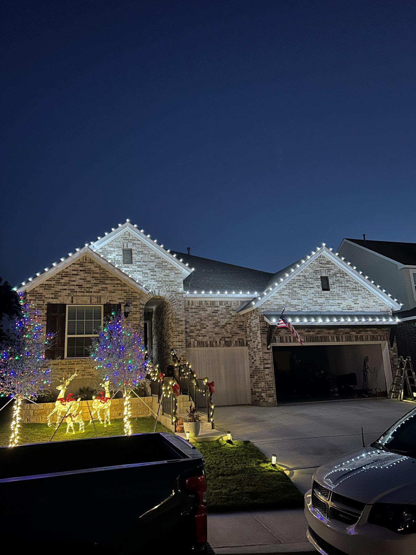 Brick home with lit reindeer, multicolor wrapped trees and garland on the stair railing